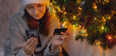 millennial woman makes online Christmas shopping, holding a mobile phone and a credit card, next to a Christmas garland
