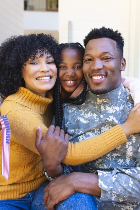Portrait of happy african american family embracing outside home with usa flag, copy space. Expression, togetherness, military, army, protection, duty, patriotism and nationality, unaltered.
